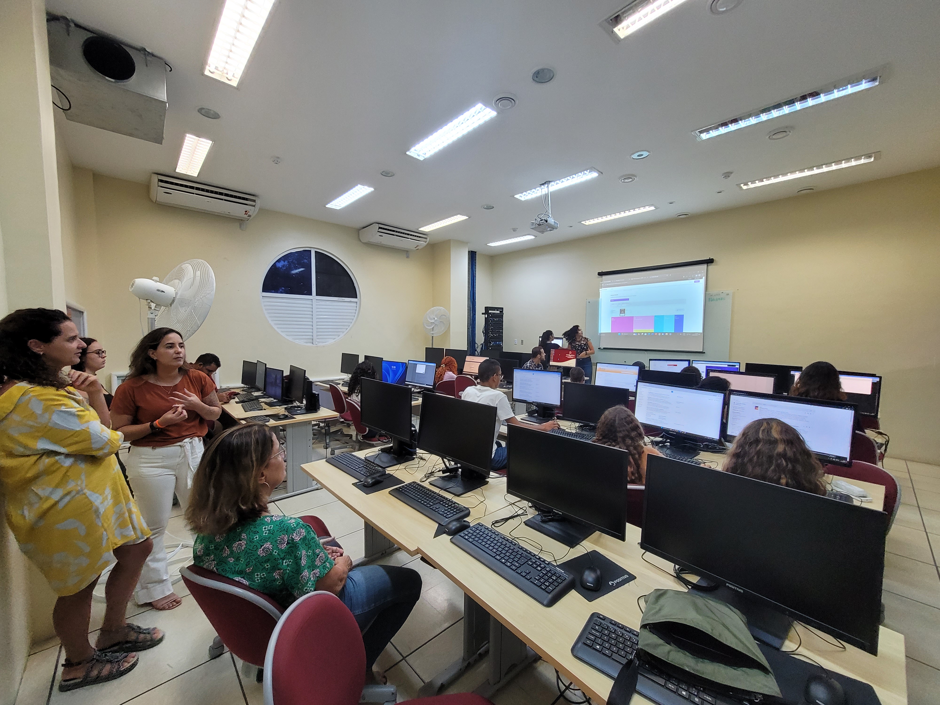 estudantes sentados de frente a computadores e os professores na frente projetando slides no quadro.